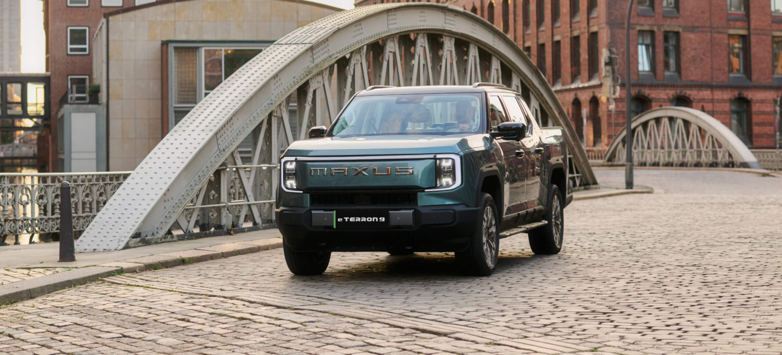 MAXUS eTERRON 9 electric pickup truck driving on a cobblestone street with a metal bridge and historic buildings in the background.